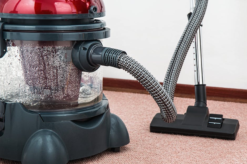 A person kneeling on a patterned carpet in a residential room, using a yellow and black vacuum cleaner with a transparent dust container to perform surface cleaning. The individual is holding a cleani