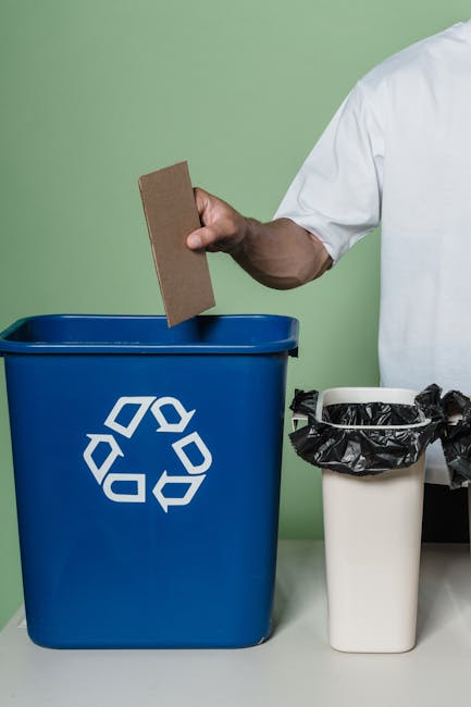 A person dressed in a blue shirt, yellow vest, and green gloves is standing outdoors on a grassy field during daytime, holding a black trash bag open with their left hand while using a white scoop or