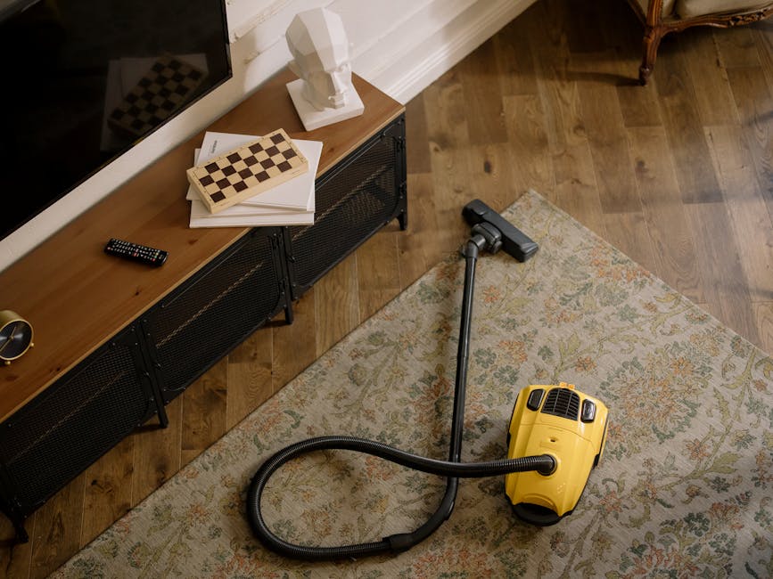 A close-up view of a black vacuum cleaner head pressing down on a detailed, patterned rug with floral motifs in muted colors. The vacuum cleaner's hose is connected to a wand held by an individual, vi