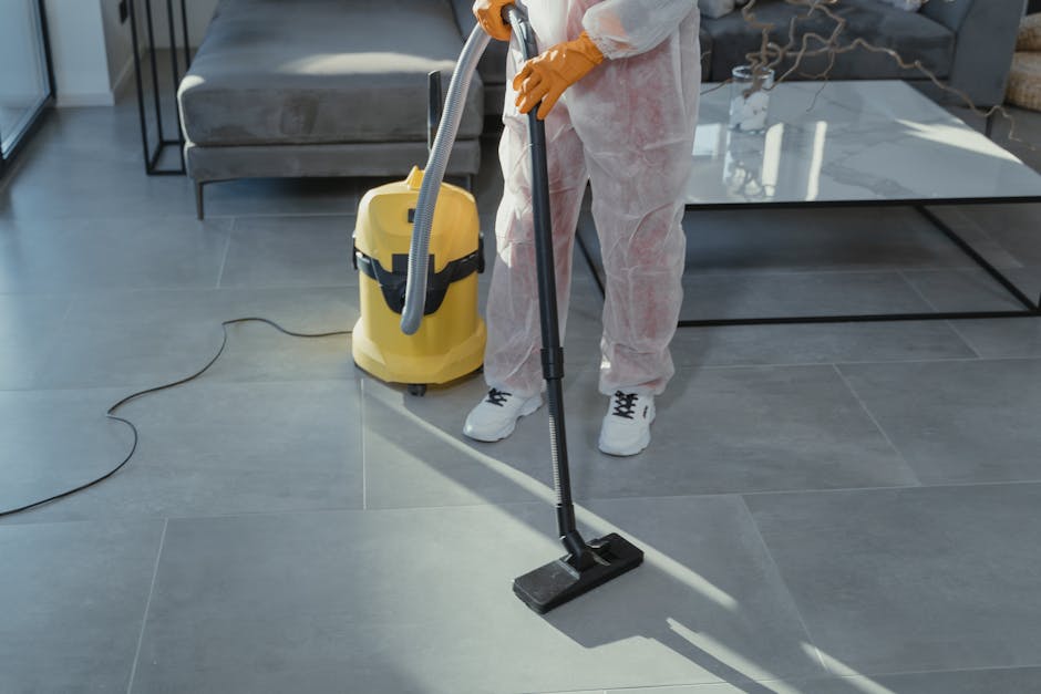 A person wearing bright pink rubber gloves is cleaning a curved, light pink ceramic sink with a green and white spray bottle and a black cloth, in a room with unfinished brick walls. The sink and surr