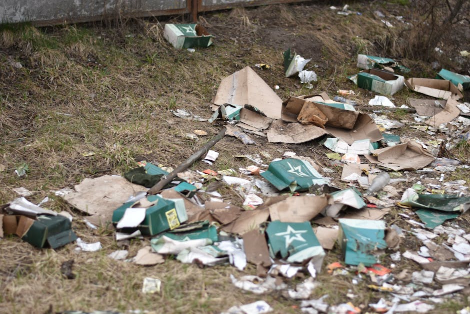 An outdoor scene showing a large pile of mixed waste and recycling bins overflowing onto the pavement, with various cardboard boxes, plastic bags, paper, and discarded packaging spilling out. Behind t